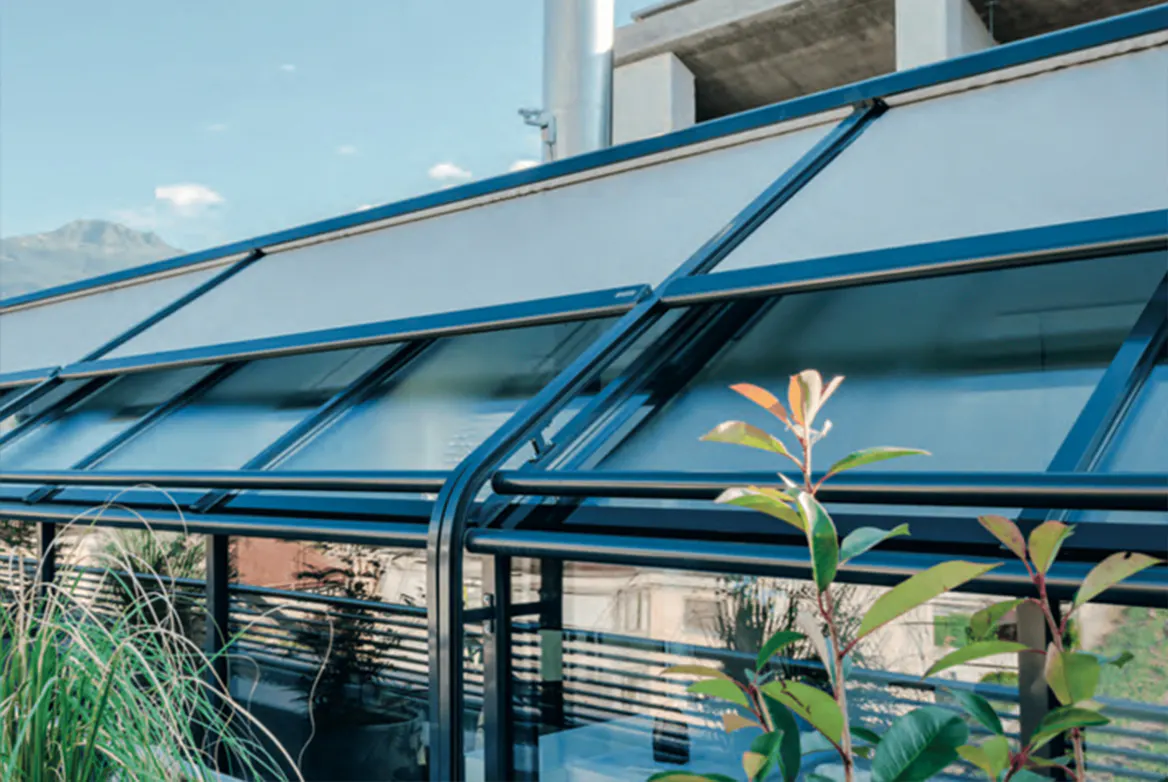 Utvendig solskjerming dekker skrå glasstakvinduer, gir skygge og beskytter mot sol på en moderne vinterhage med blåmalte metallrammer, planter i forgrunnen og bygninger under blå himmel.