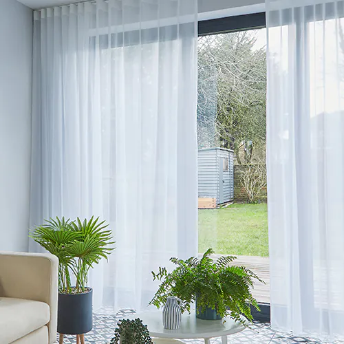 Gjennomsiktige, hvite gardiner henger foran skyvedør og filtrerer dagslys i en lys stue med terrasse, gressplen og et lite hageskur; potteplanter står foran vinduet.