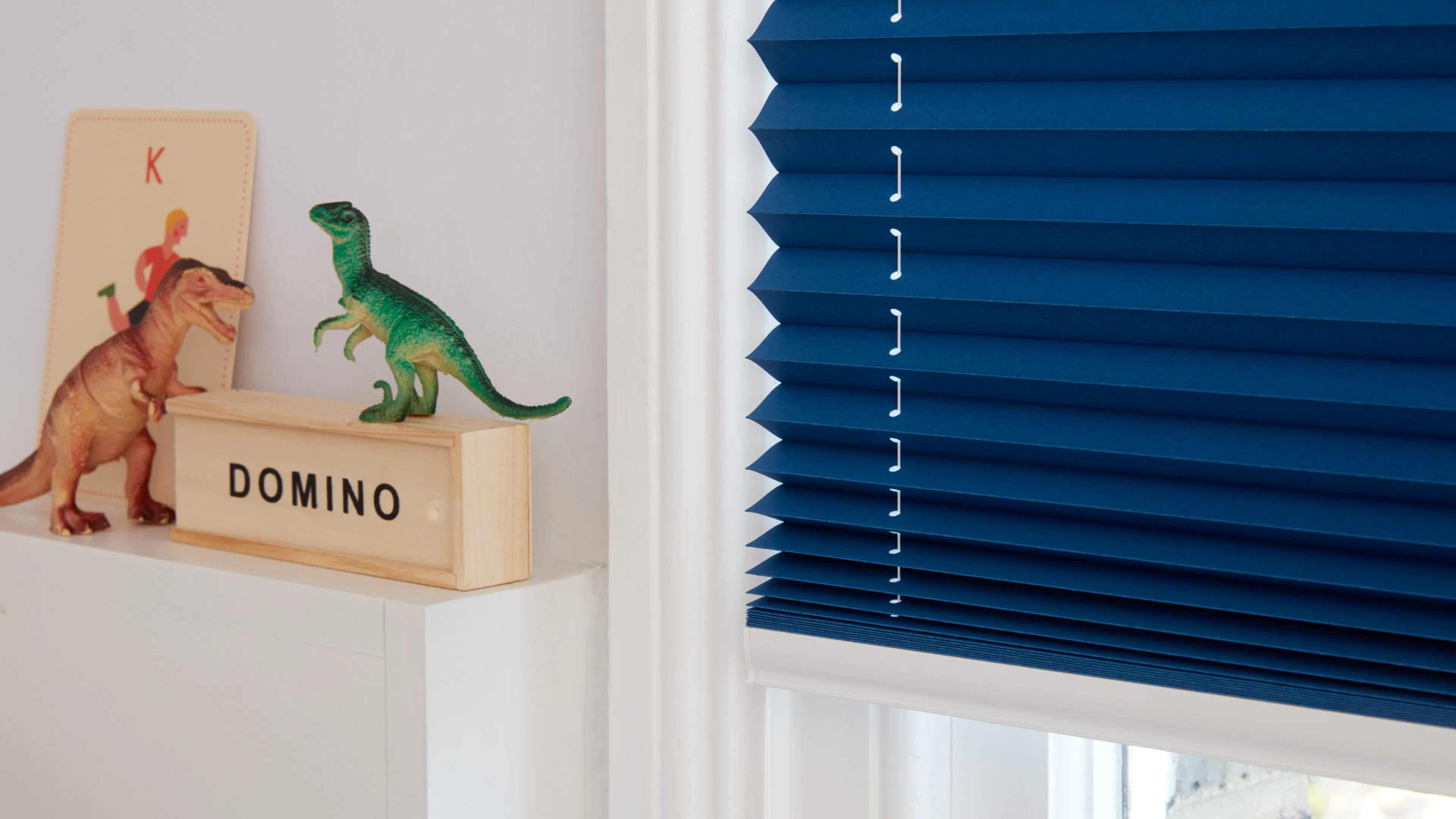 Blå plisségardin er delvis trukket ned foran et hvitt vindu. På en hylle ved vinduskarmen står en treskuff merket "DOMINO" med en grønn dinosaurleke.