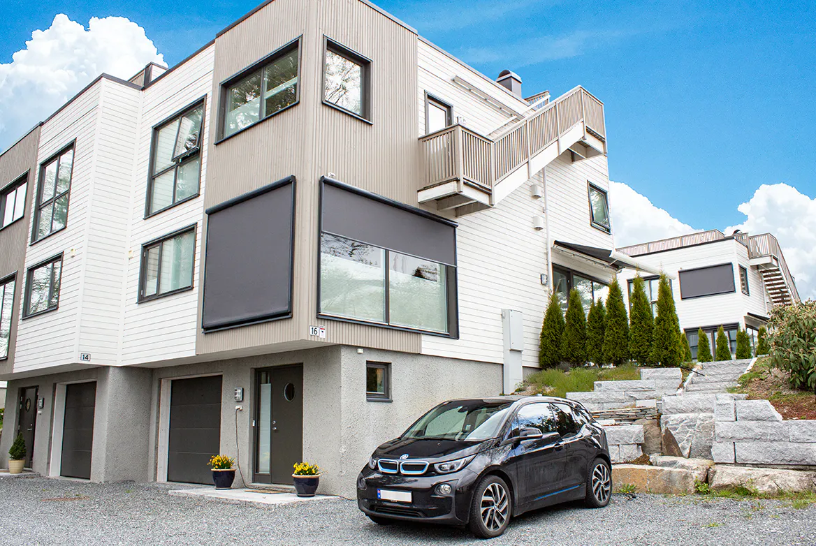 Utvendige rullegardiner dekker store vinduer på et moderne hvitt og grått hjørnehus. En svart elbil står parkert i oppkjørselen ved steintrapp og hekk under klar blå himmel.