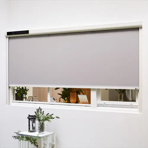 Rullgardin dekker et vindu delvis; lys grå rullegardin henger i en hvit kasse og gir privatliv mens nederste glassrute viser grønne planter, med et hvitt bord, lykt og vase foran.