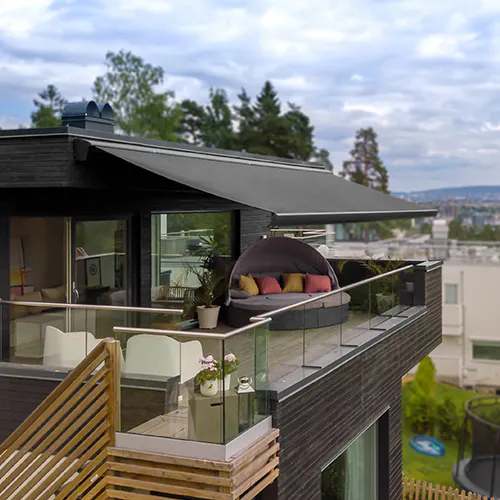 Utvendig terrassemarkise dekker en sittegruppe på en moderne tre- og flislagt uteplass, beskytter inngang og skyvedører; glassrekkverk, planter og nabohus vises i bakgrunnen.