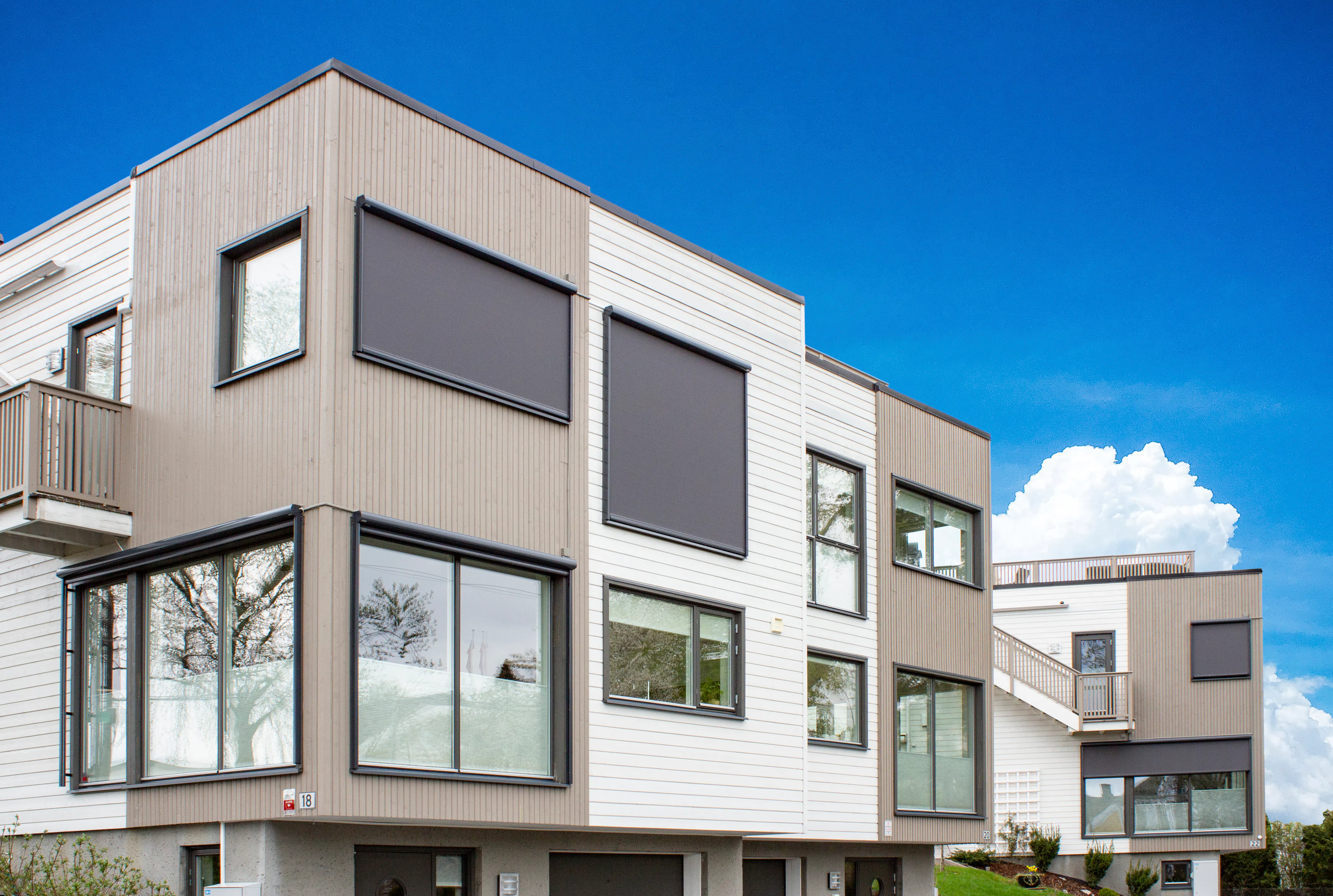 Utvendige grå rullegardiner dekker flere store vinduer på en moderne, lys, tre- og panelkledd leilighetsbygning under klar blå himmel med gressplen foran.