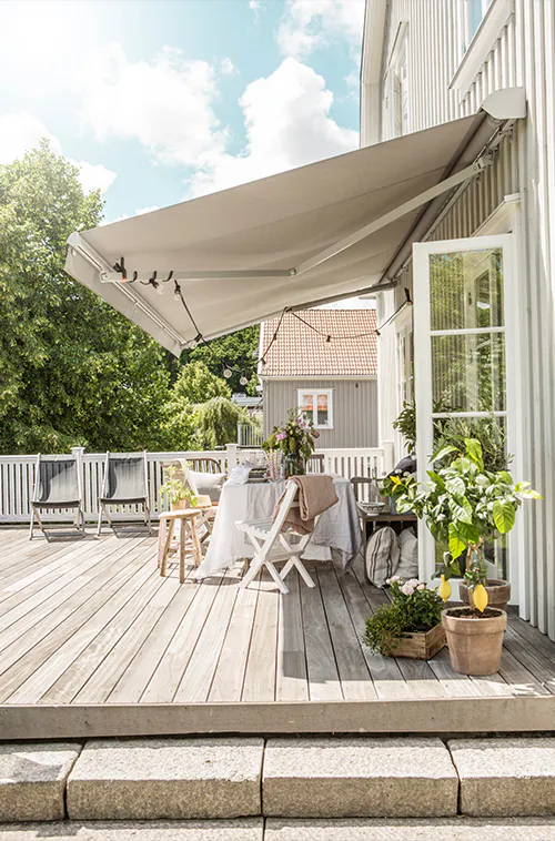 Markise er trukket ut over en solrik treterrasse; et bord dekket for utendørsmåltid, stoler, planter og åpne terrassedører danner en koselig hagekontekst mot hvitt rekkverk og nabohus.