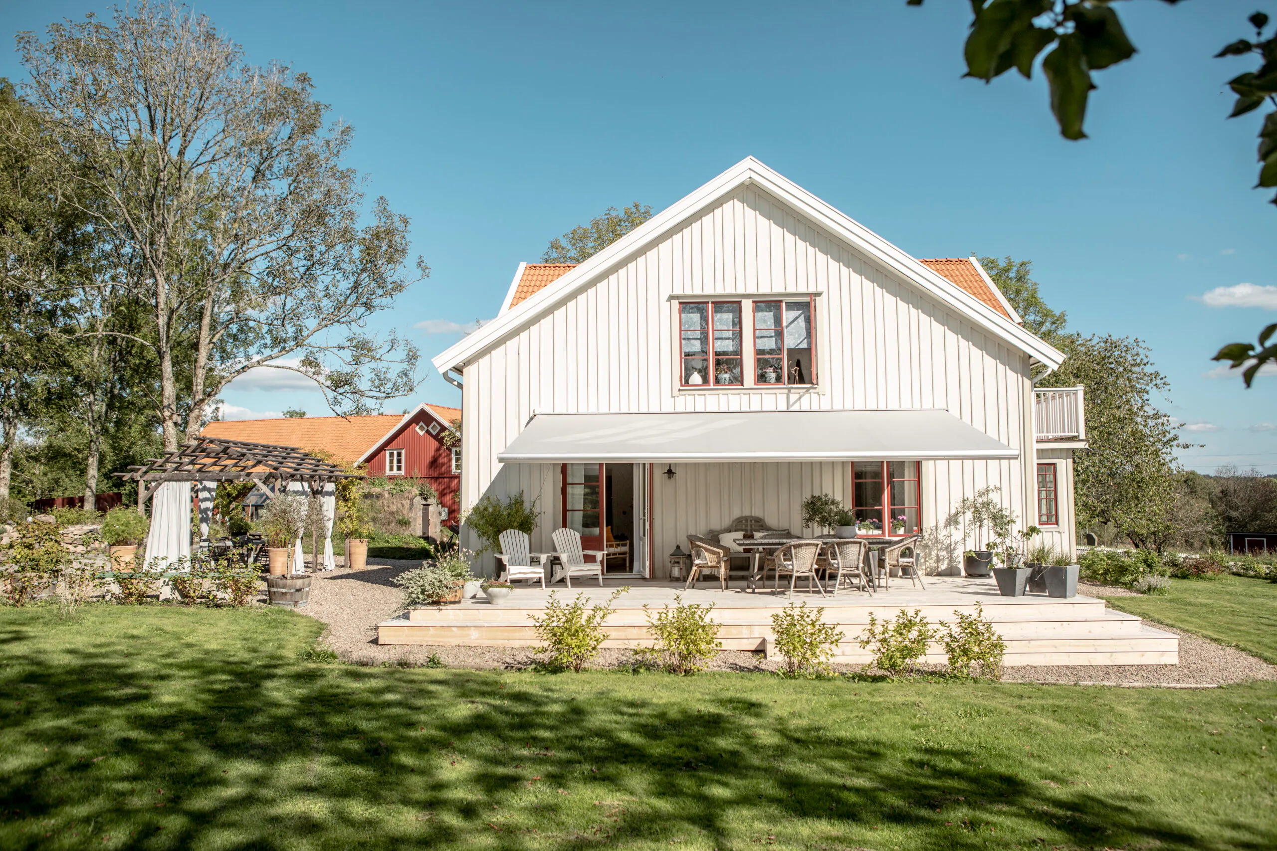 Hvite utegardiner henger delvis trukket til side i en overbygd terrasse og rammer inn glassdører; huset står i en solrik hage med grønn plen, busker, trær og ute møbler.