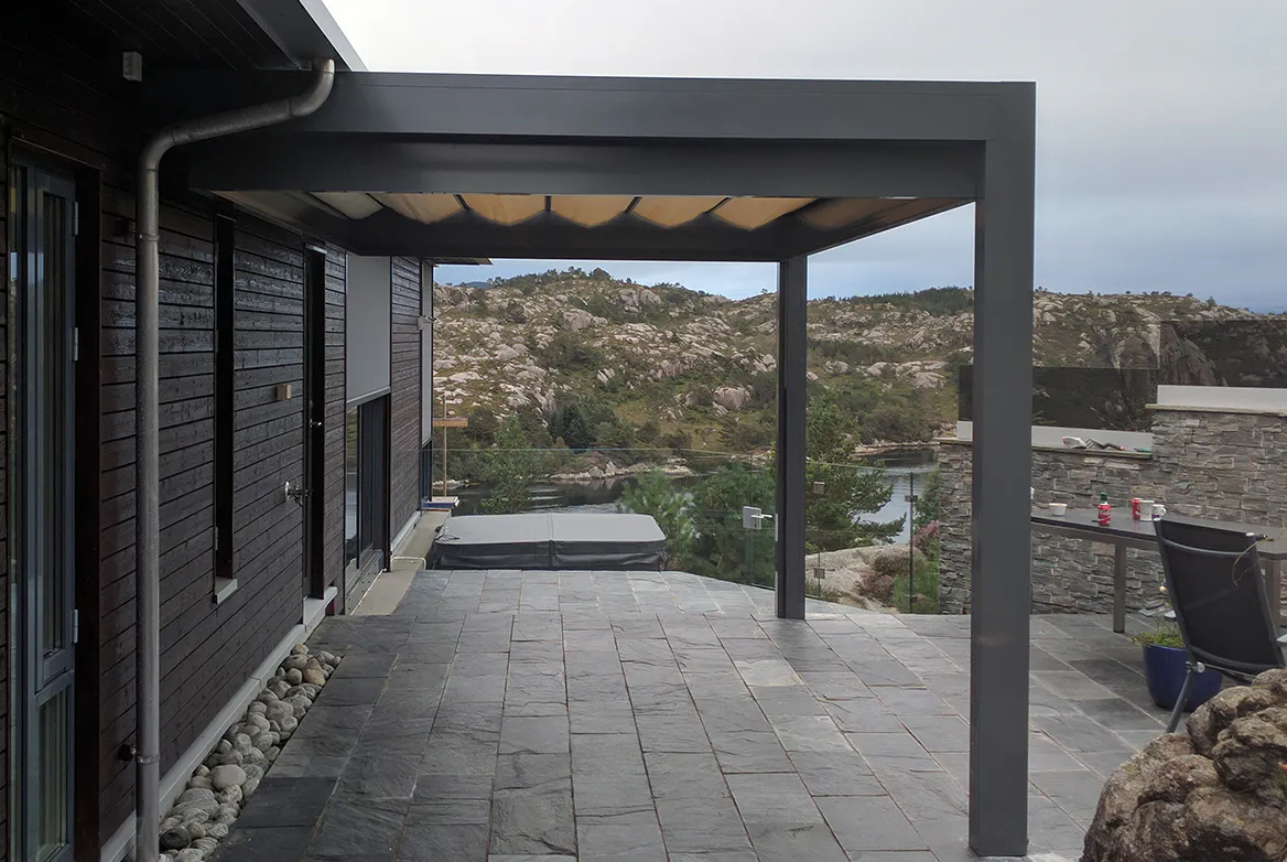 Pergola med nedtrekkbar stoffmarkise som skygger over steinlagt terrasse; glassrekkverk, boblebad og utebord i et moderne husmiljø med utsikt mot klippefylt kystlandskap.