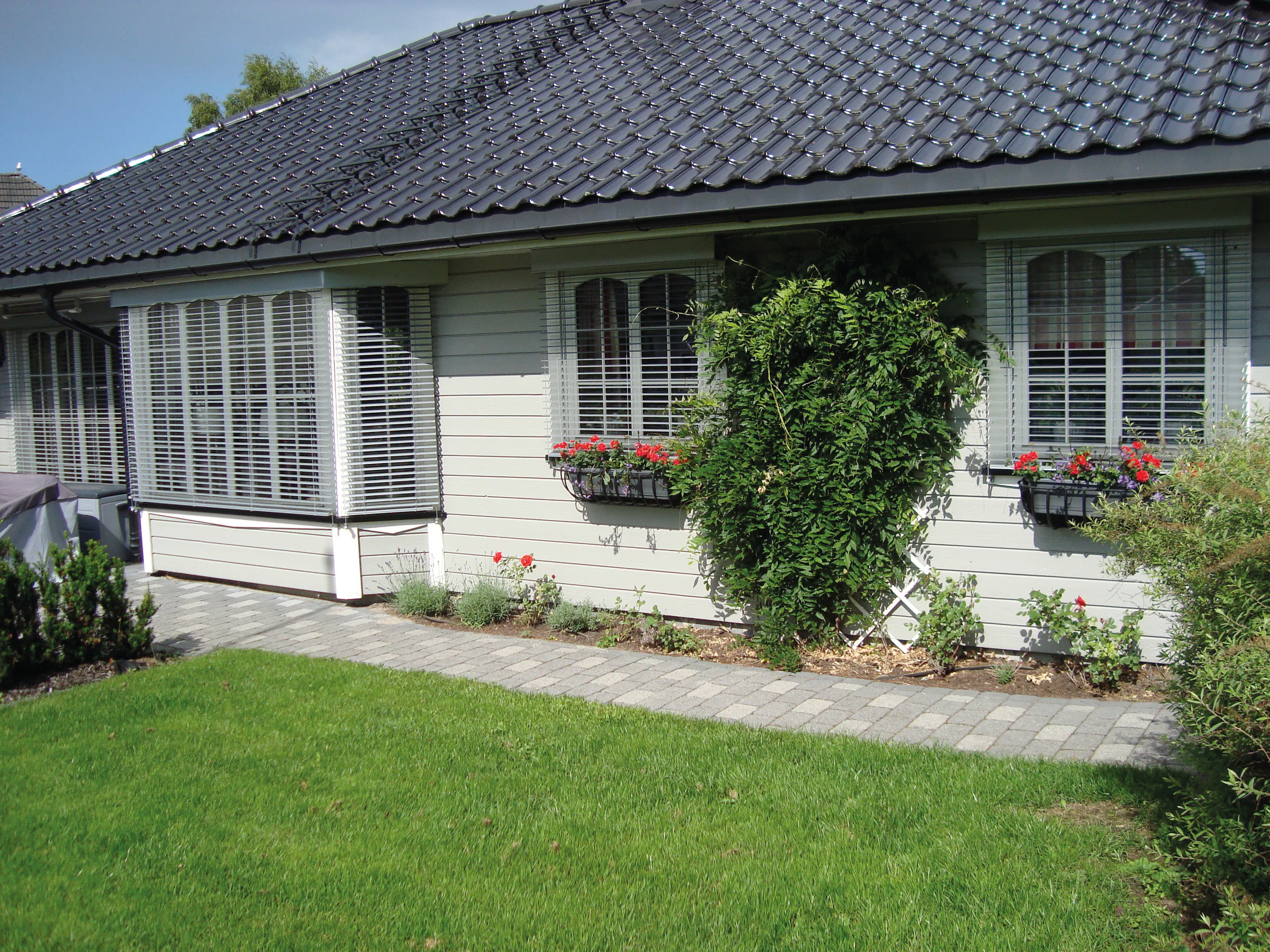 Hvite utvendige skodder dekker flere brede vinduer på en enetasjes trevilla; blomsterkasser under vinduene, brosteinssti langs huset og velstelt grønn plen i forhaven.