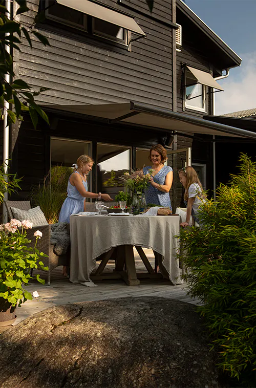 En mørk markise gir skygge over en uteplass; tre kvinner dekker et bord med duk og blomster foran et moderne, svart trehus, omgitt av frodig grønn beplantning.