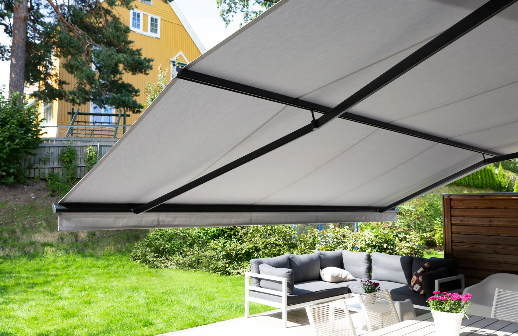 En utfelt grå markise strekker seg ut over en terrasse; den skygger en sittegruppe med grå puter ved et hvitt bord, med grønn plen og et gult hus i bakgrunnen.
