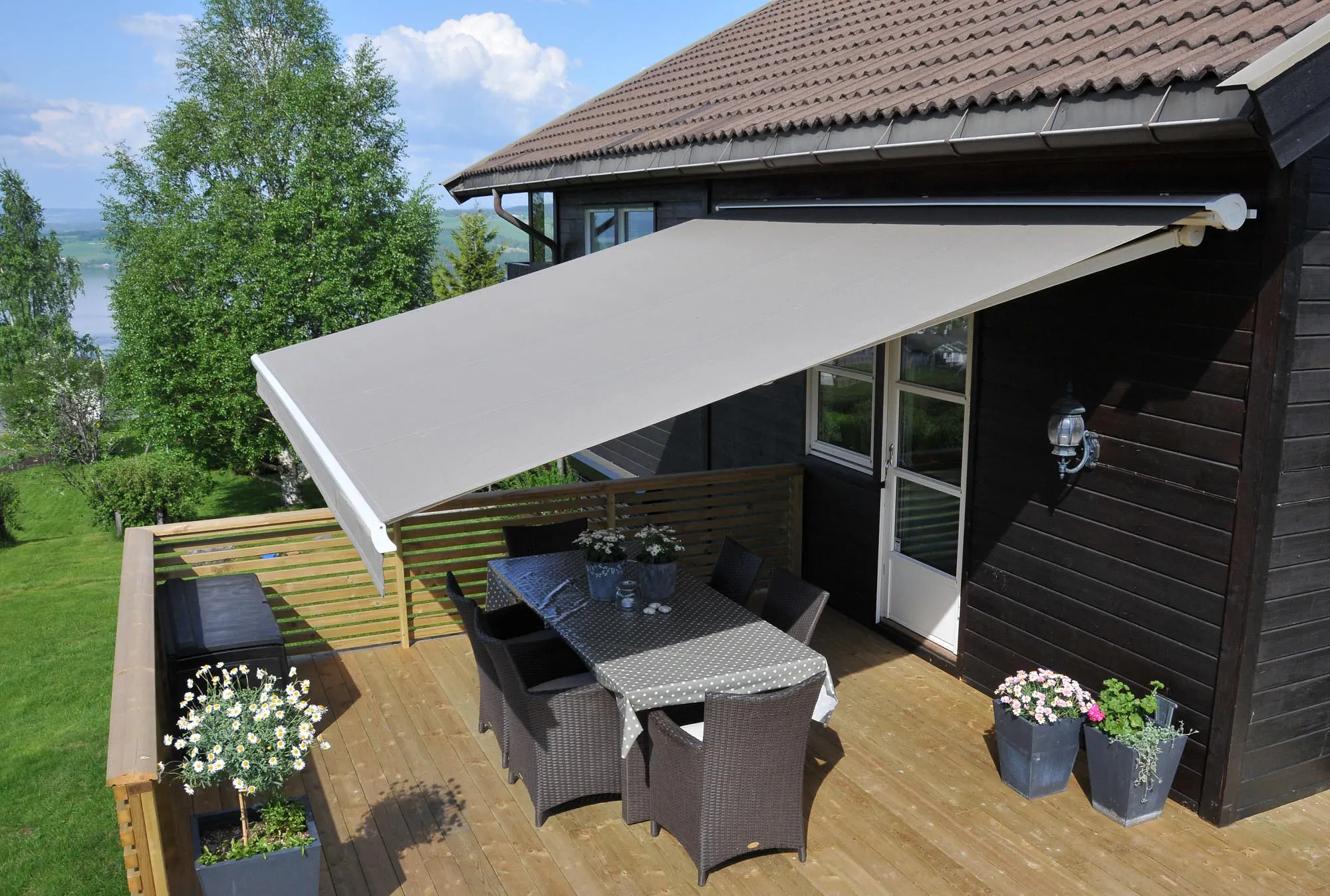 En lys grå uttrekkbar markise dekker en solrik treterrasse og gir skygge over en spisegruppe med fire kurvstoler; husfasade, blomsterpotter og utsikt mot innsjø og trær i bakgrunnen.