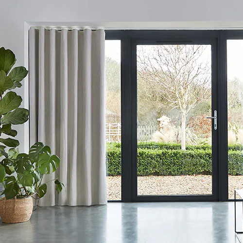 Lys grå gardin henger til venstre av store svarte terrassedører, delvis trukket til side; glass viser en beskjært hekk og hage; polert betonggulv og potteplante til venstre.
