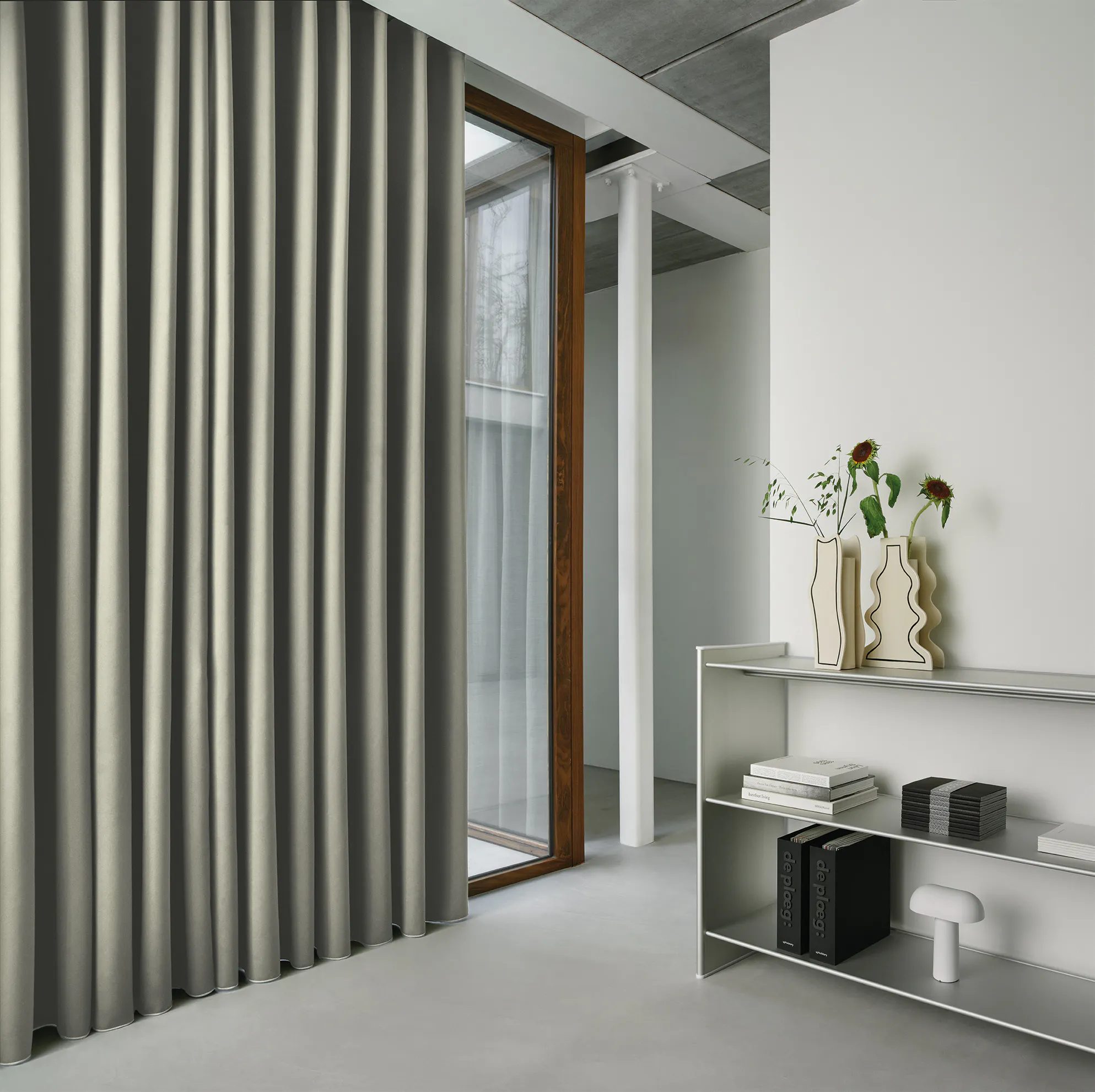 Mørkegrå, tunge gardiner henger delvis for et stort glassdørvindu; rommet er lyst og minimalistisk med hvit vegg, metallhylle med bøker, dekorative vaser og en synlig søyle.