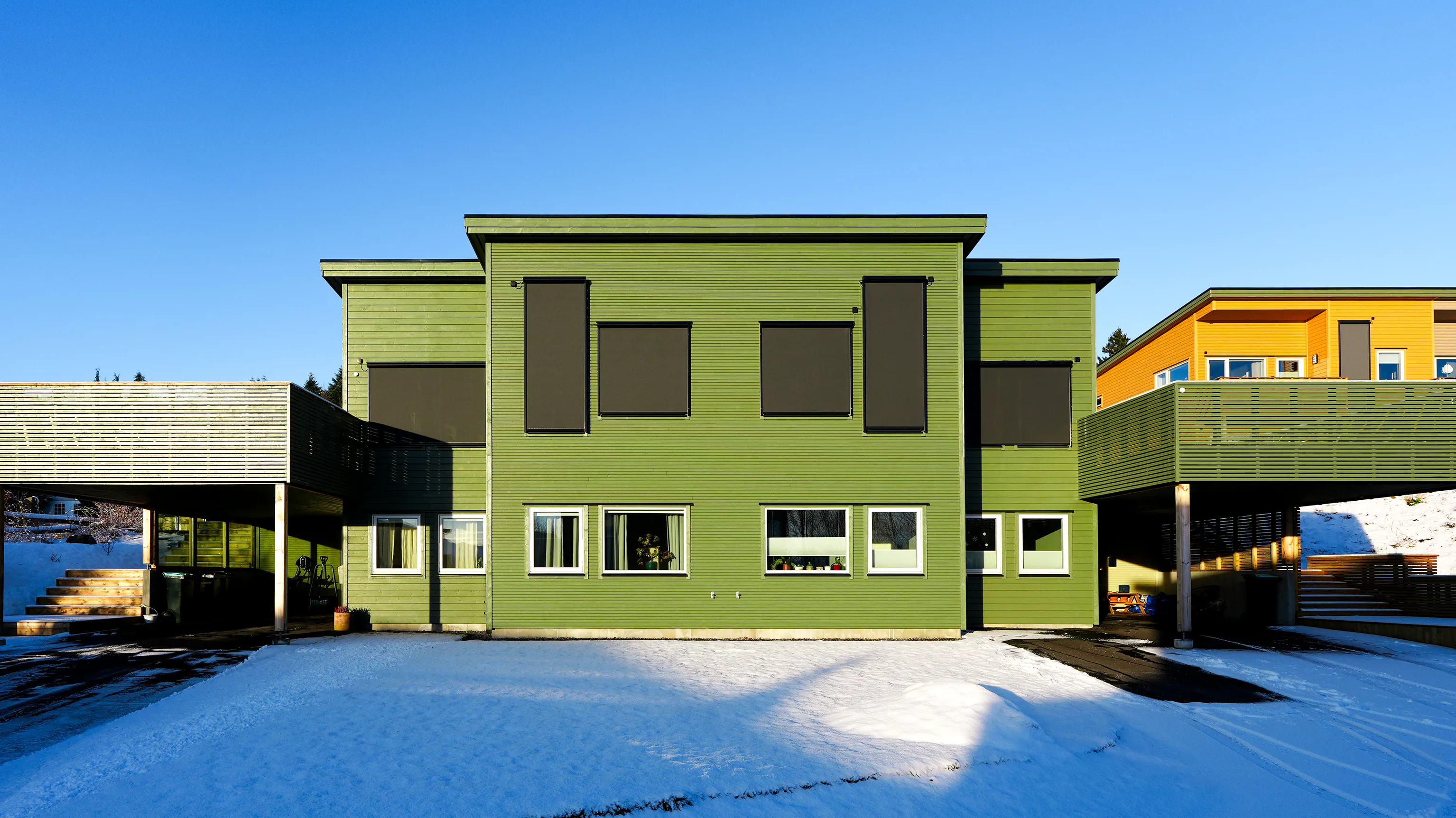 Grønt moderne bygg med mørke utvendige rullegardiner trukket ned over øvre vinduer; tre nederste vinduer ubeskyttet. Snødekt forplass og klar blå himmel i bakgrunnen.