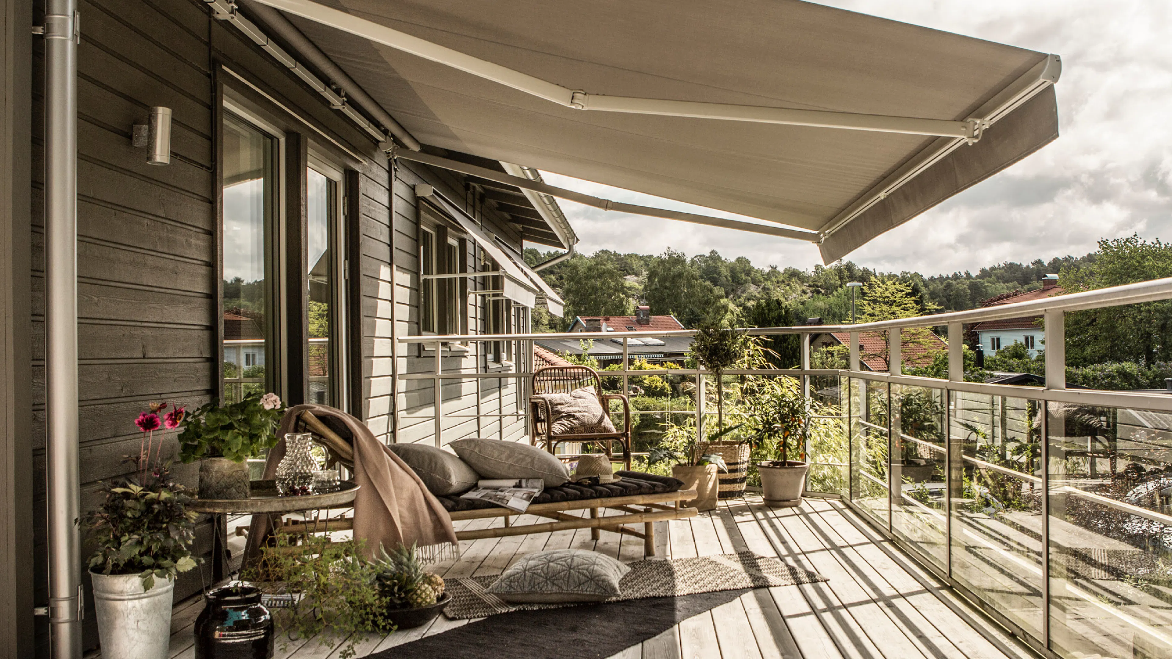 En kremfarget terrassemarkise er trukket ut og gir skygge over en møblert balkong med solsenger, puter og planter; glassrekkverk og grønne trær danner en rolig forstadskontekst.