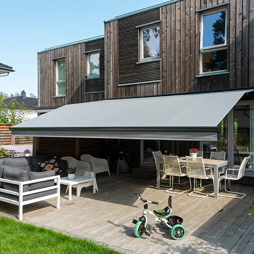 Stor uttrekkbar markise dekker en terrasse med bord og stoler, skygger for sitteområde foran en to-etasjes bolig med vinduer og balkong.