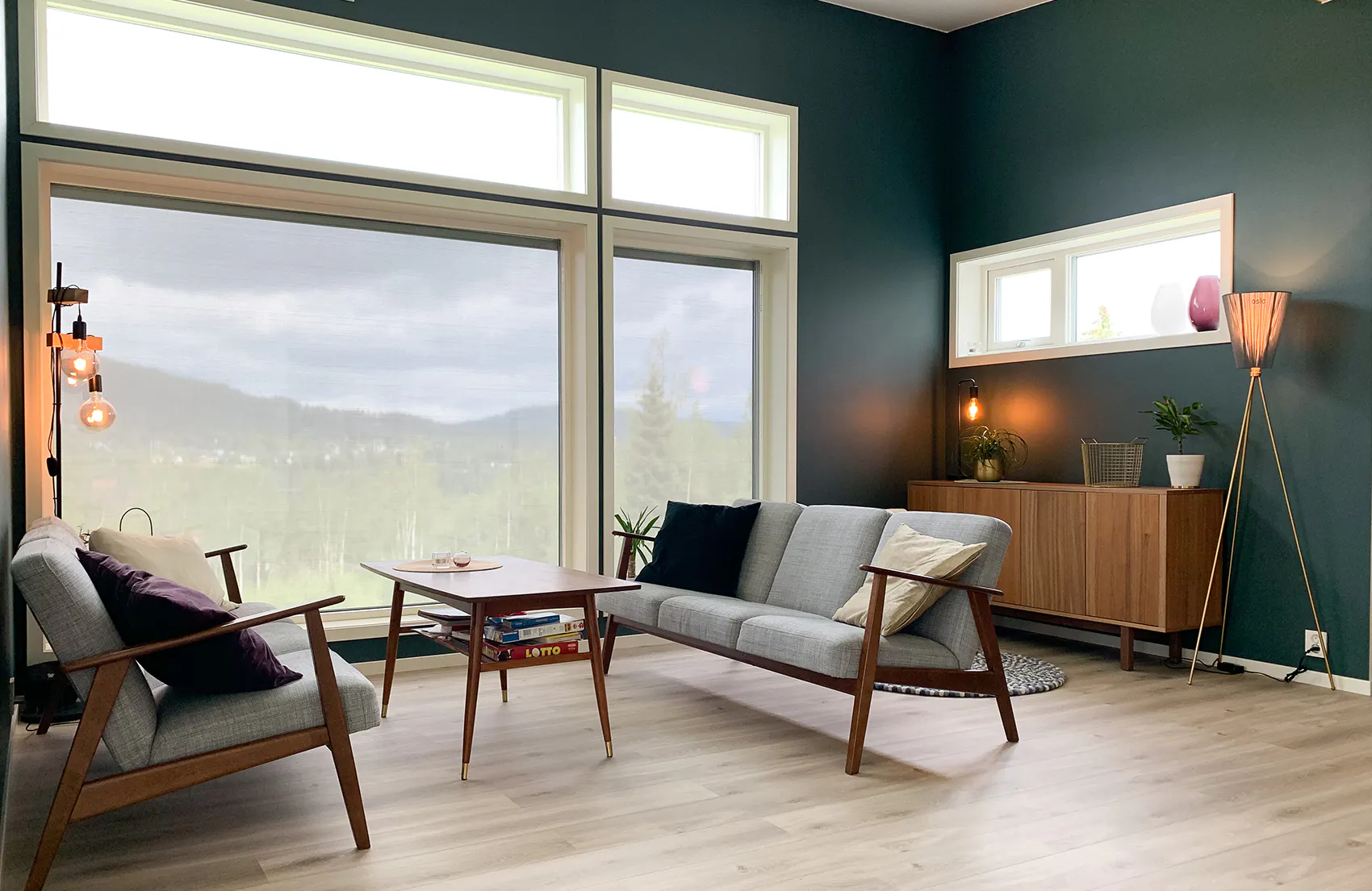 Store, nedrullede lysfiltrerende rullegardiner dekker store vinduer, demper innslipp av lys i en moderne stue med sofaer, trebord, grønn vegg, sideboard, gulvlampe og utsikt mot åser.