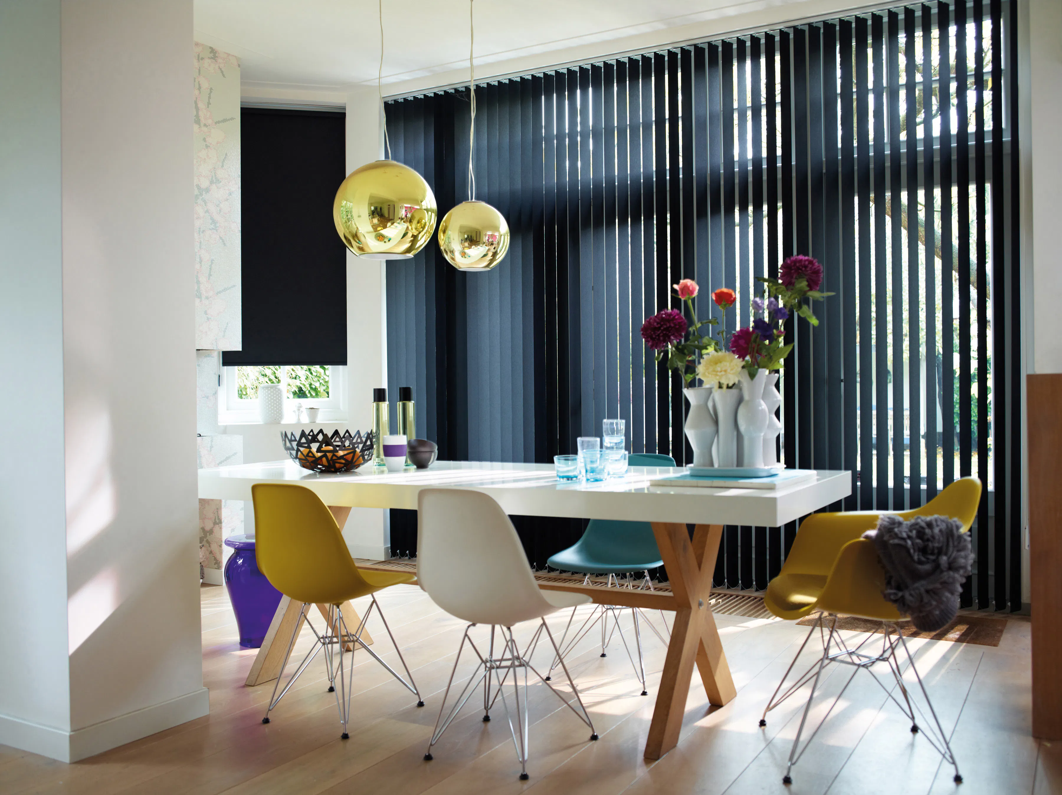 Vertikale lamellgardiner dekker høye vinduer og regulerer lys i en moderne spisestue med hvitt bord, fargerike designstoler, hengende gullpendler og en vase med blomster.