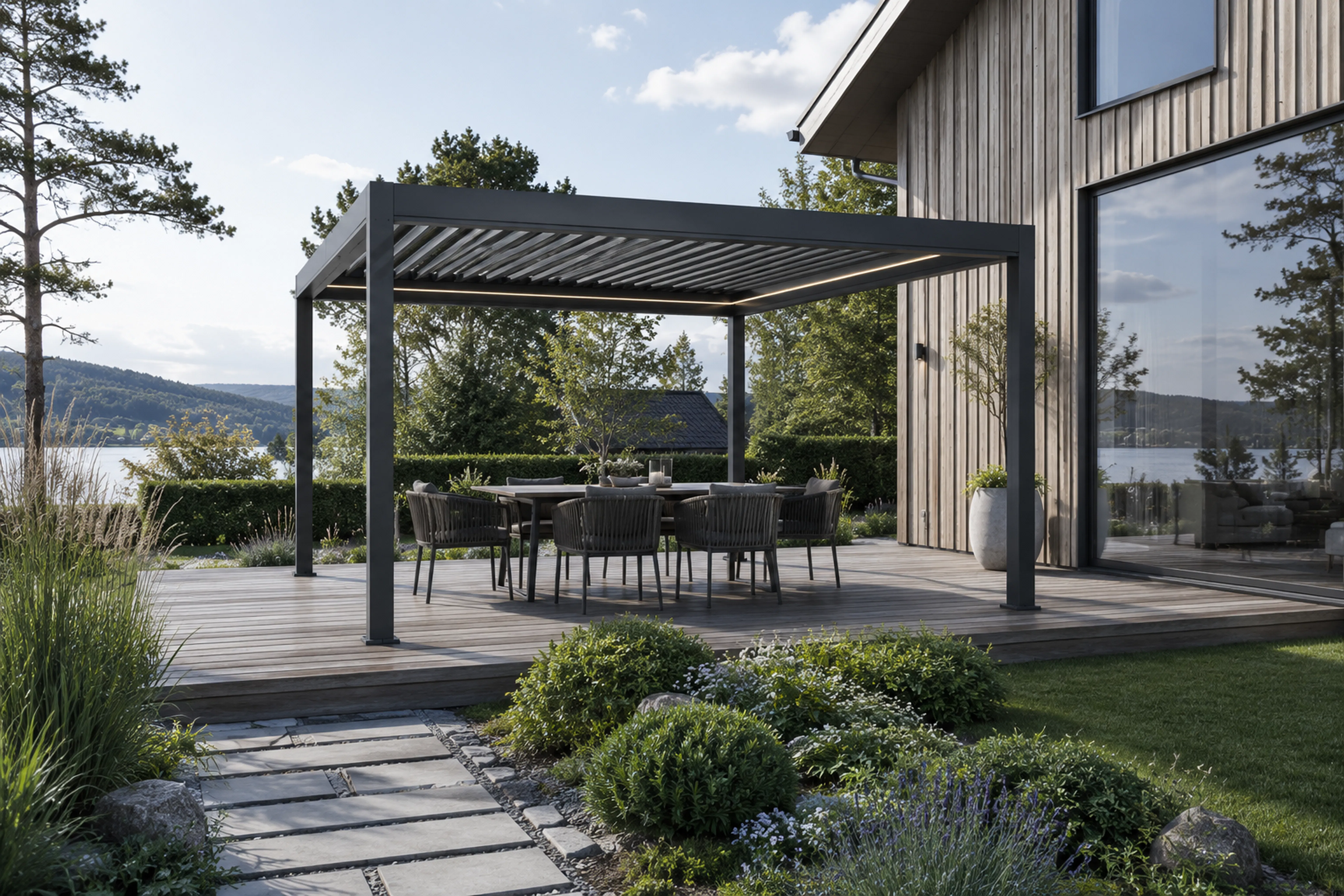 Mørkegrå pergola med justerbare lameller skaper skygge over et utespisebord og stoler på en treterrasse ved en moderne husfasade, omgitt av hage, busker og innsjøutsikt.