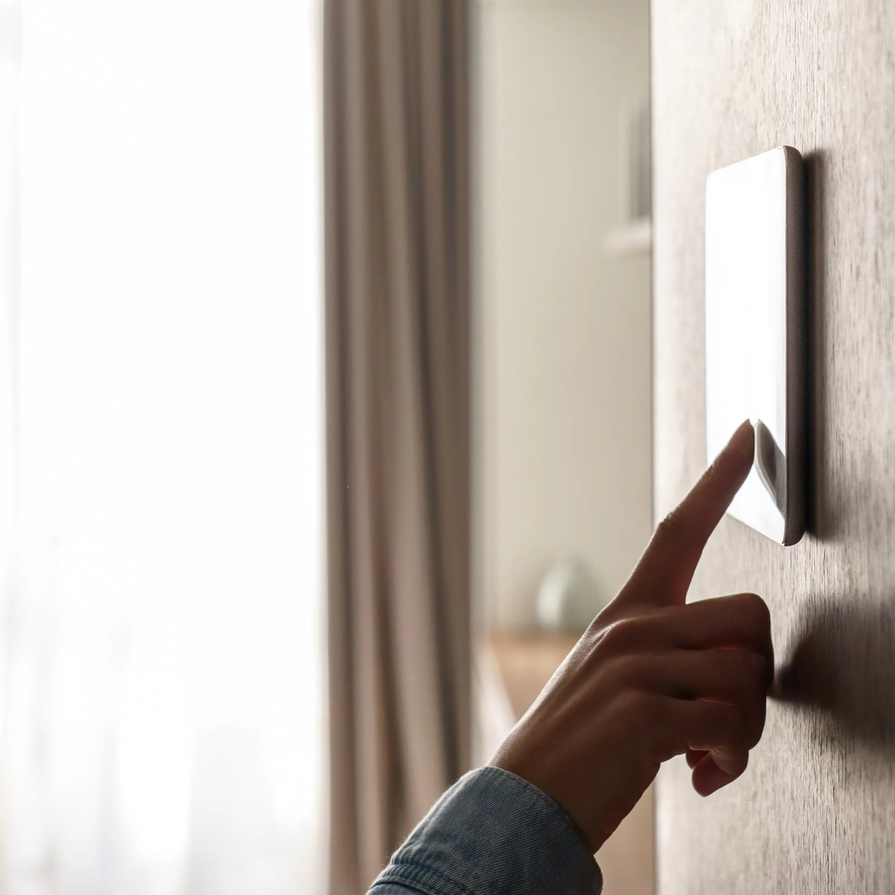 En beige gardin henger delvis foran et lyst vindu, mens en finger trykker en moderne veggbryter på en teksturert vegg i et dempet, moderne soveromsinteriør.