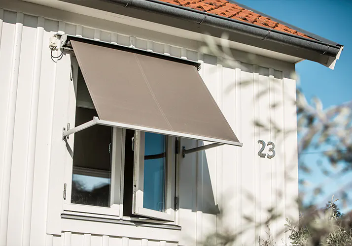 Et vindu utstyrt med grønn-hvit stripete markise som er trukket ut, gir skygge over et åpent sidehengslet vindu på en lys, delvis mønstret husfasade.