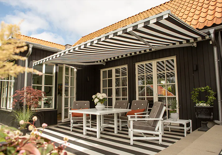 Stripete sorthvitt markise gir skygge over terrasse foran et svart trehus med hvite vinduer; matchende stripete terrassegulv, hvitt hagebord og stoler, potteplanter og rødt tak.