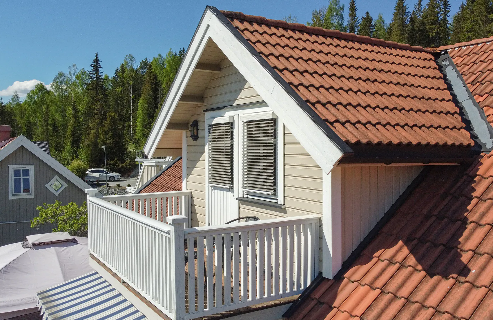 Hvite horisontale persienner dekker et loftsvindu i en kvist, er delvis lukket for å skjerme lys. Hvit balkongrekkverk, rødt tegltak og furuskog i bakgrunnen.