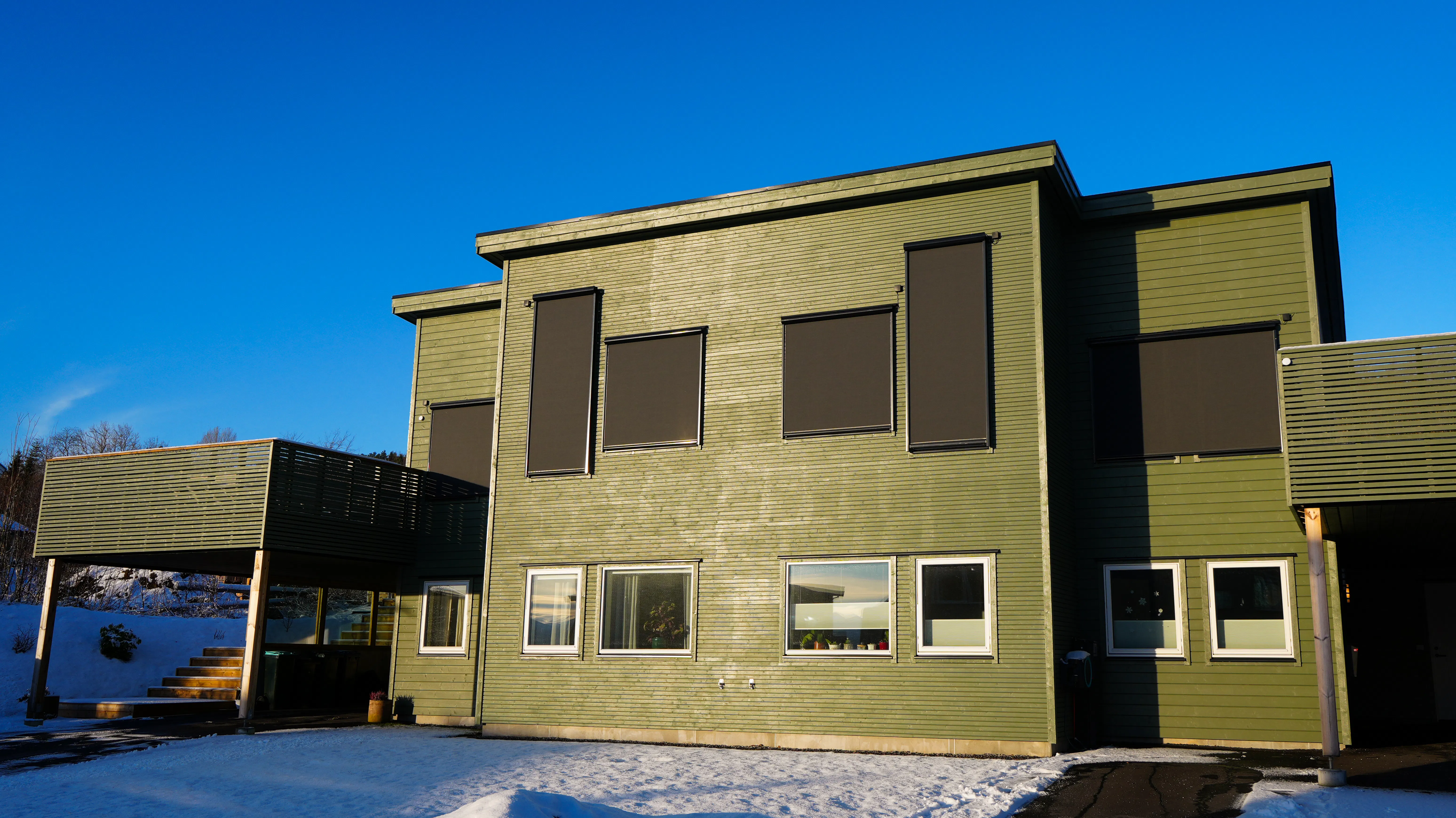 Utvendige mørke rullegardiner dekker flere vinduer på en grønn trekledning; huset står i lavt sollys ved snødekt forhage under en klar blå himmel.