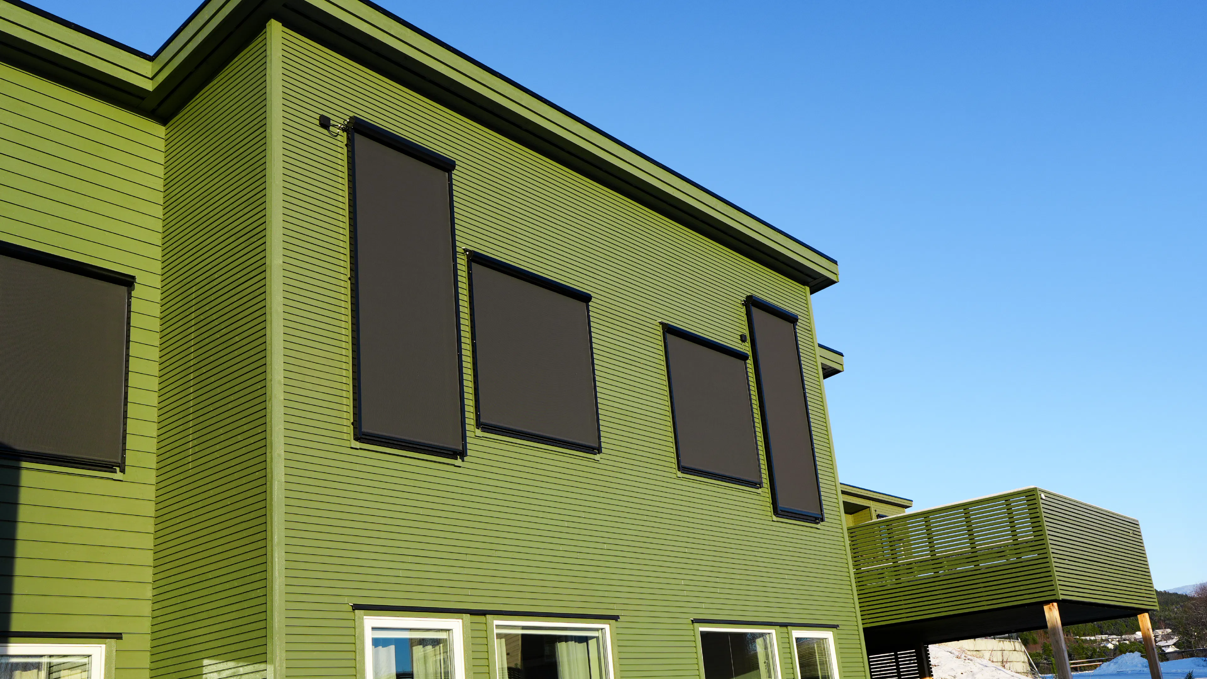 Mørke utvendige rullegardiner dekker flere vinduer på en grønn to-etasjes bygning; et balkongrekkverk vises, huset står i sterkt sollys under en klar blå himmel.