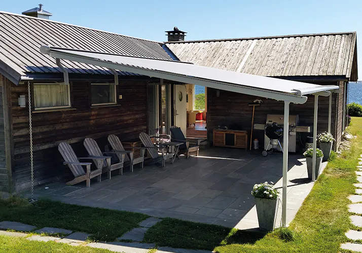 En hvit, uttrekkbar markise dekker en steinlagt terrasse, gir skygge over utemøbler (fire trestoler, bord), grill og oppbevaringskasse ved en mørk trehusfasade under blå himmel.