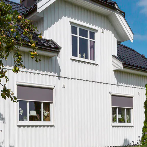 To grå rullegardiner er delvis trukket ned i de to nederste vinduene på en hvit trekledd villa; øvre vindu har gardin, og et epletre med frukt står til venstre.