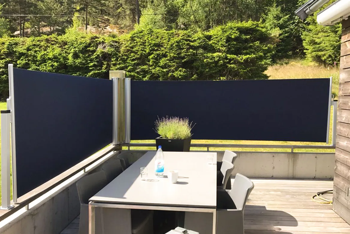 Mørkeblå balkongskjerm dekker rekkverk og avskjermer utsikten; et bord med to stoler, en plante og en sprayflaske står på tredekke, med grønn hekk og skog i bakgrunnen.