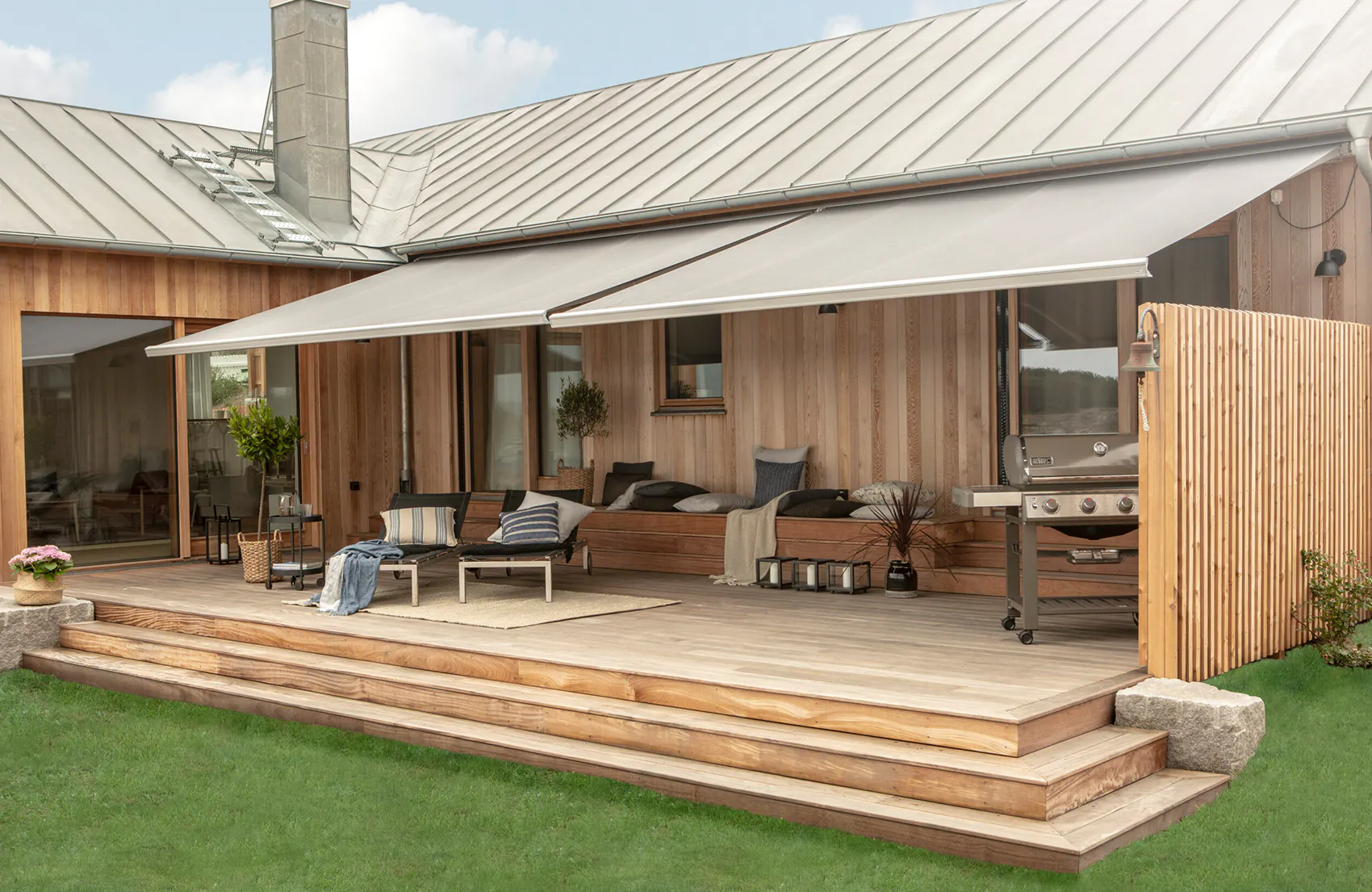 Uttrekkbar markise gir skygge over en treplatting med sofagruppe, puter og sofabord; terrasse ved et moderne trehus, trappetrinn ned til plen, gassgrill ved høyre ende.