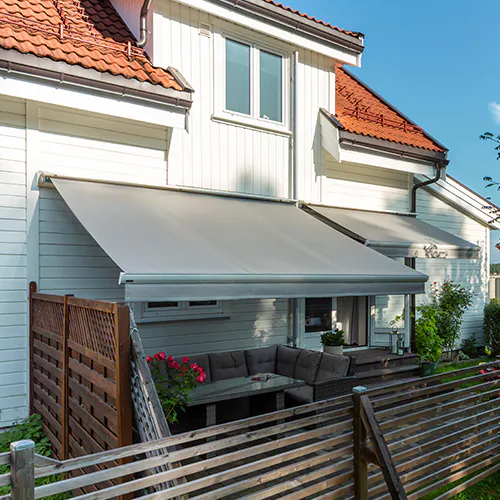 En utvendig grå markise er trukket ut over en terrasse, gir skygge til hagemøbler ved et hvitt trehus med røde takstein og et tregjerde i solskinn.