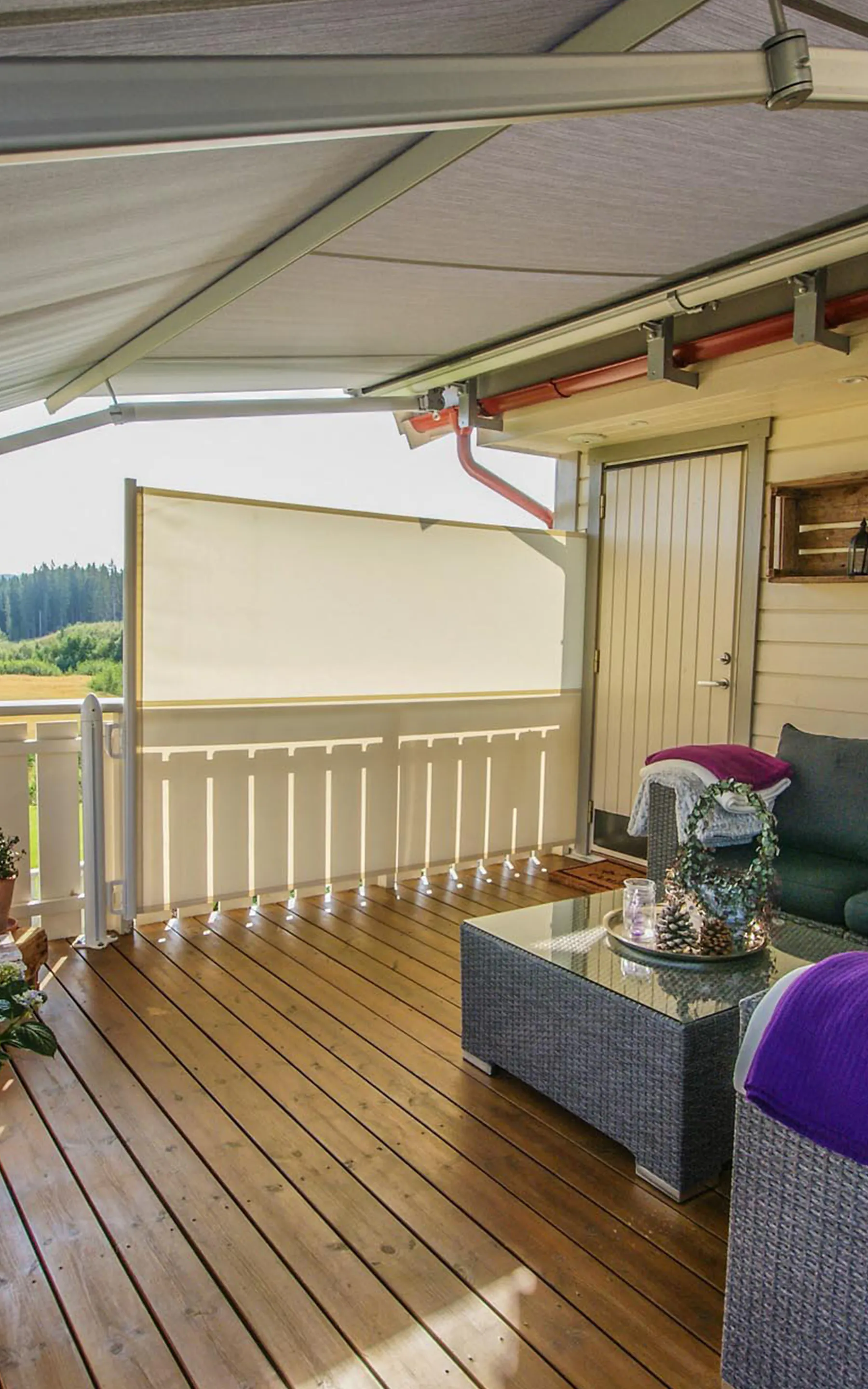 En utvendig rullegardin henger ned og skygger en liten overbygd terrasse; den demper sollys og gir privatliv mens tredekke, potteplanter, en sofa med lilla pute og en inngangsdør omgir området.
