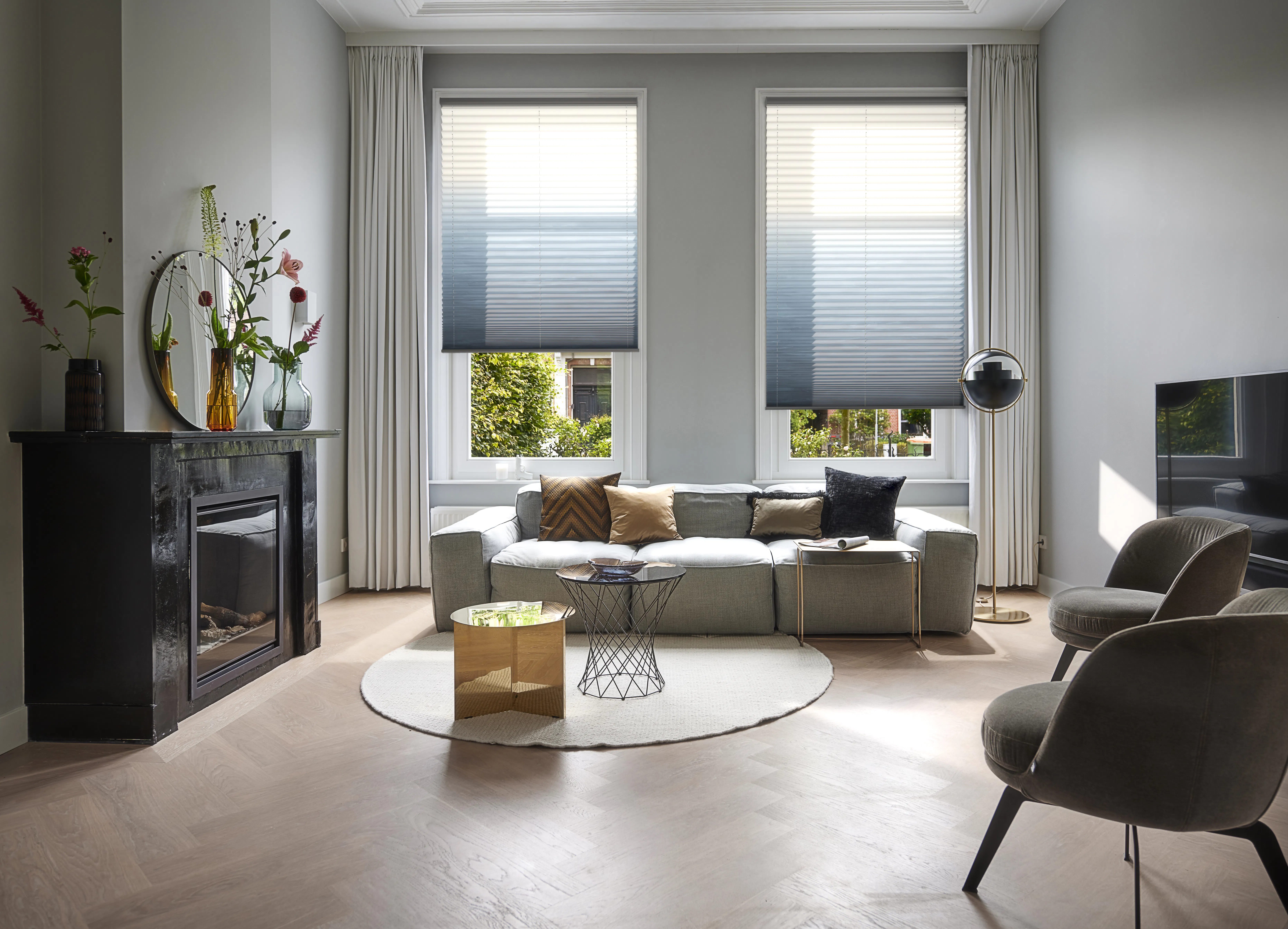 To grå-hvite plisségardiner er delvis senket foran to sidestilte vinduer; innrammet av lange hvite gardiner i en lys, moderne stue med sofa, peis, bord og dekor.