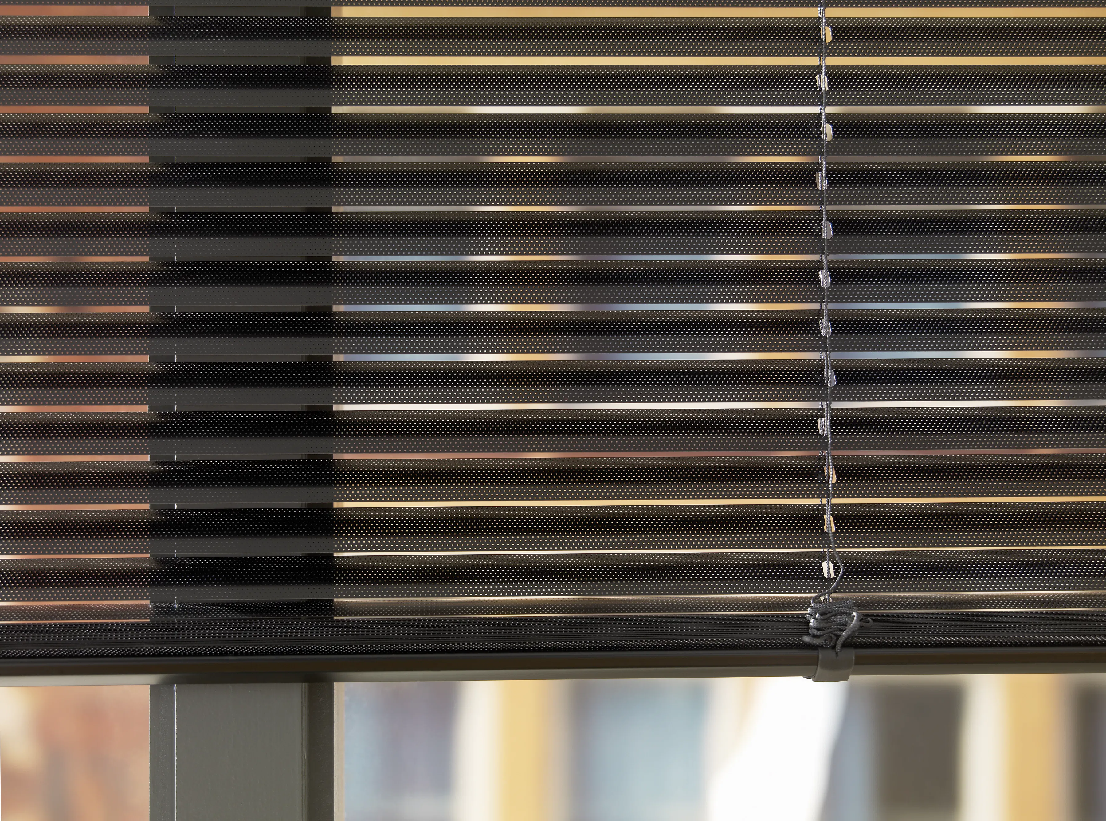 Persienne i mørkegrå aluminiumslatser delvis lukket; slattenes horisontale linjer demper sollys mens tau og kjede henger til høyre, foran et vindu med uklart bybilde utenfor.