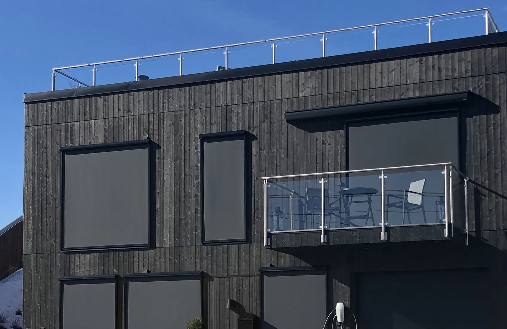 Utvendige rullegardiner dekker flere vinduer på en moderne, mørk trefasade; en balkong med glassrekkverk og hagemøbler står til høyre, under klar blå himmel.