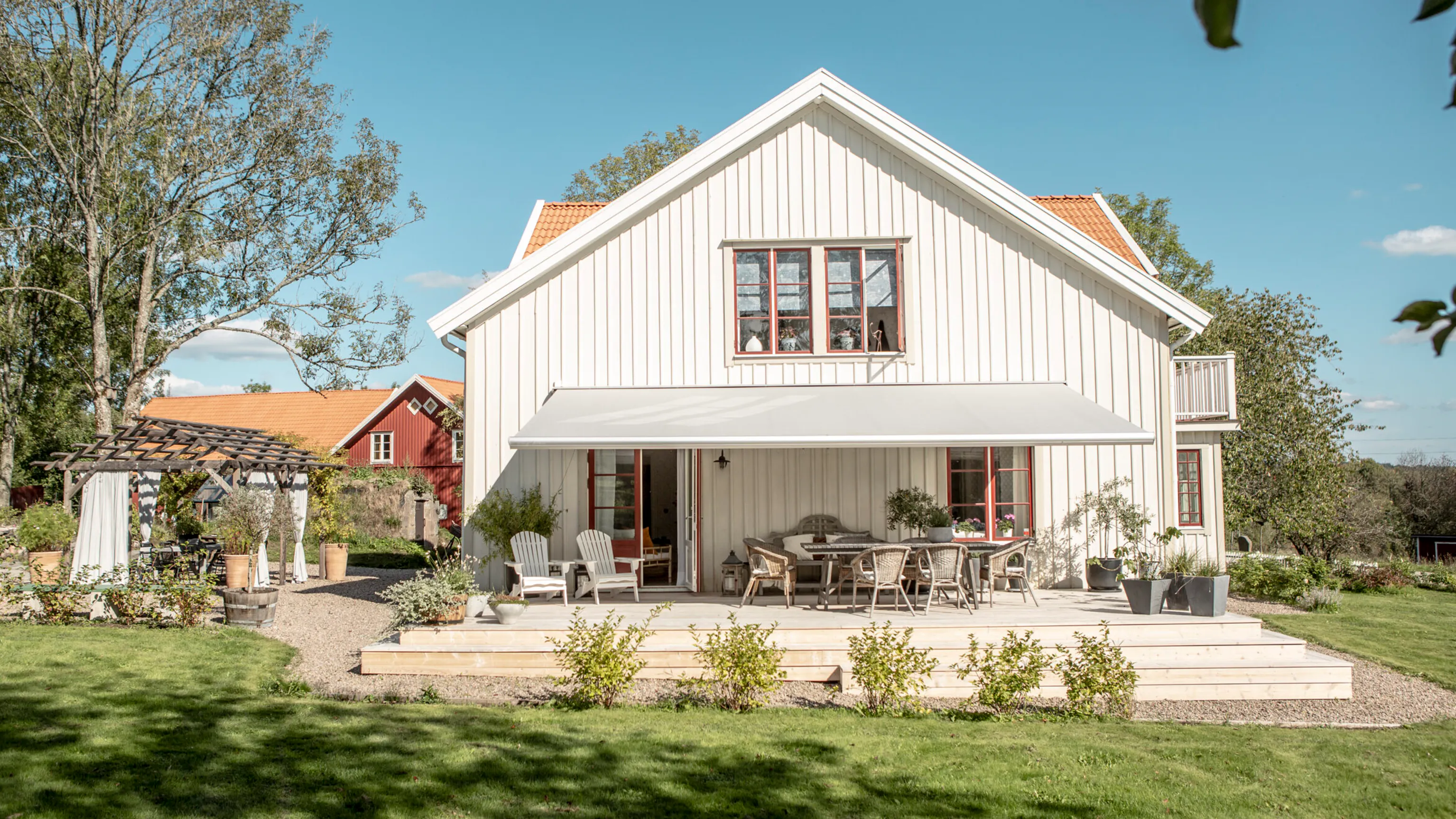 En hvit markise dekker en solrik treterrasse, skaper skygge over utemøbler og planter foran et hvitt toetasjes hus med røde vinduskarmer og grønn plen.