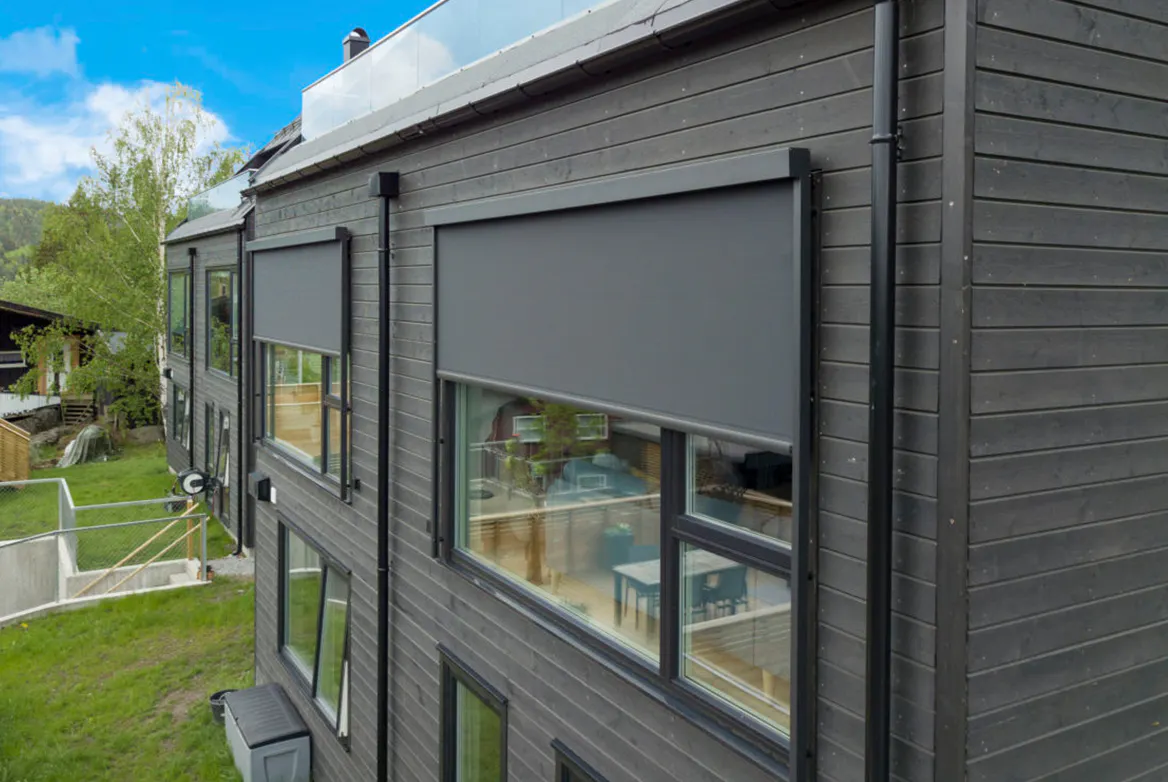 To mørkegrå utvendige rullegardiner dekker store vinduer på en moderne mørk trekledning fasade, montert over andre etasje, gir skygge og privatliv på en solrik dag.