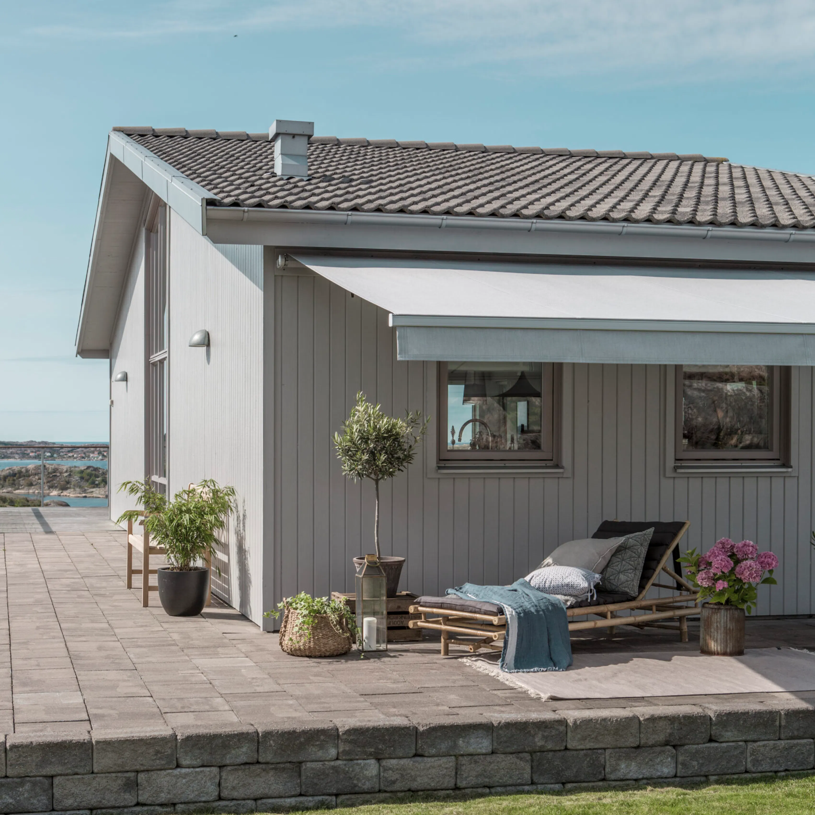 En utvendig markise/solskjerming dekker terrassen og vinduene på et grått, trepanelert sjøhus, og skygger en solseng omgitt av puter, tepper og potteplanter på flislagt uteområde.