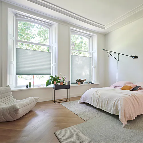 Hvite gardiner og persienner filtrerer dagslys i et lyst soverom; dobbeltseng med lyse sengetepper, nattbord med lampe og potteplante ved vinduet.