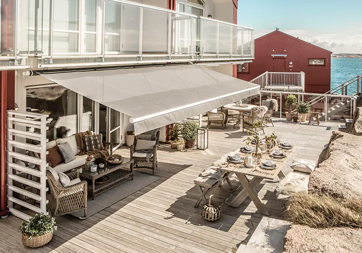 Markise foldes ut og gir skygge over en treterrasse med sittegruppe i rotting og et langbord dekket for måltid; glassrekkverk, planter og et rødt hus i bakgrunnen.