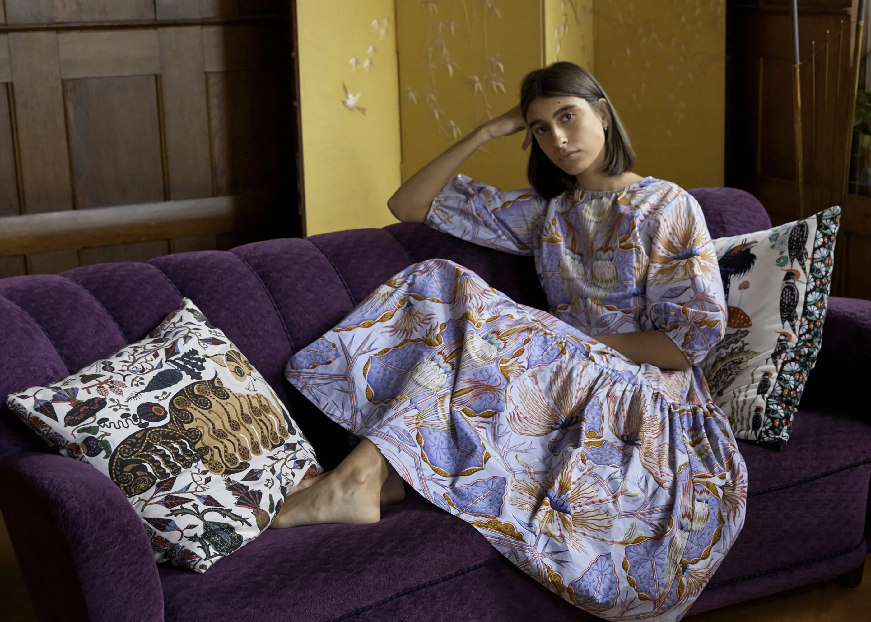 Woman in a printed dress resting on a purple velvet sofa with printed Klaus Haapaniemi cushions