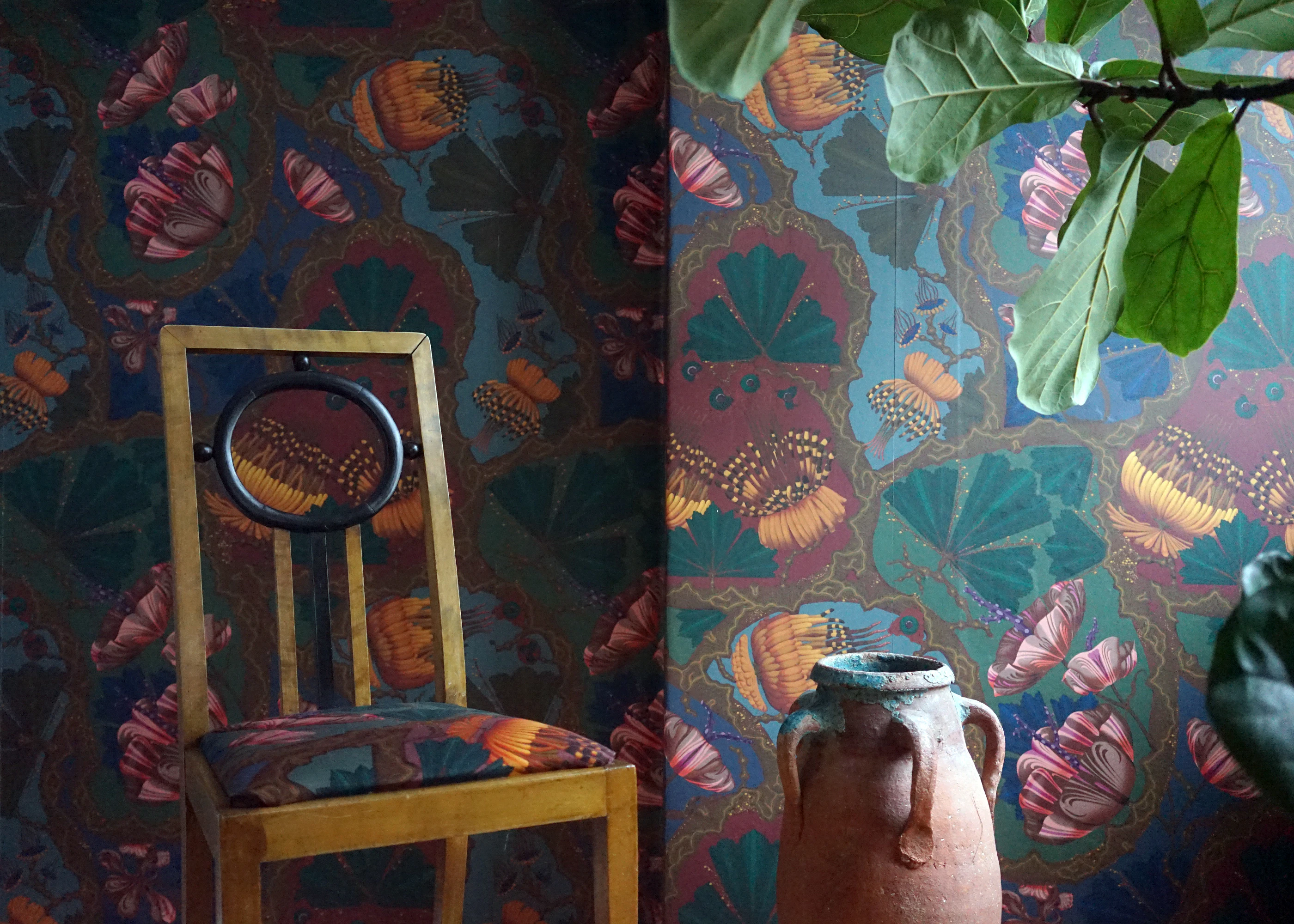 Blue Artemisia wallpaper by Klaus Haapaniemi with large folk-style flowers, behind a wooden chair with a printed seat and a terracotta urn