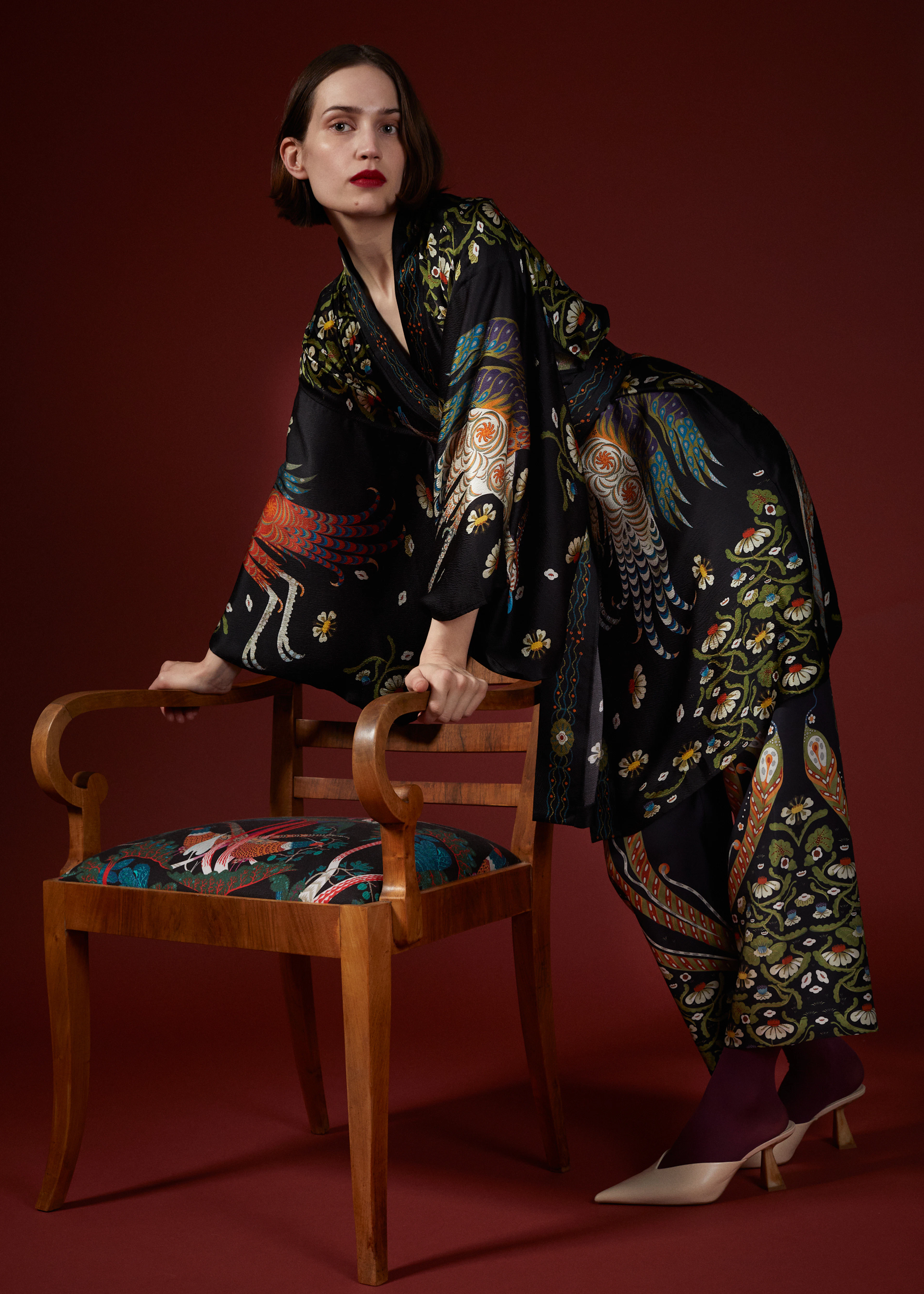 Woman in the black Firebird kimono and matching printed trousers leaning on a wooden chair against a deep red wall