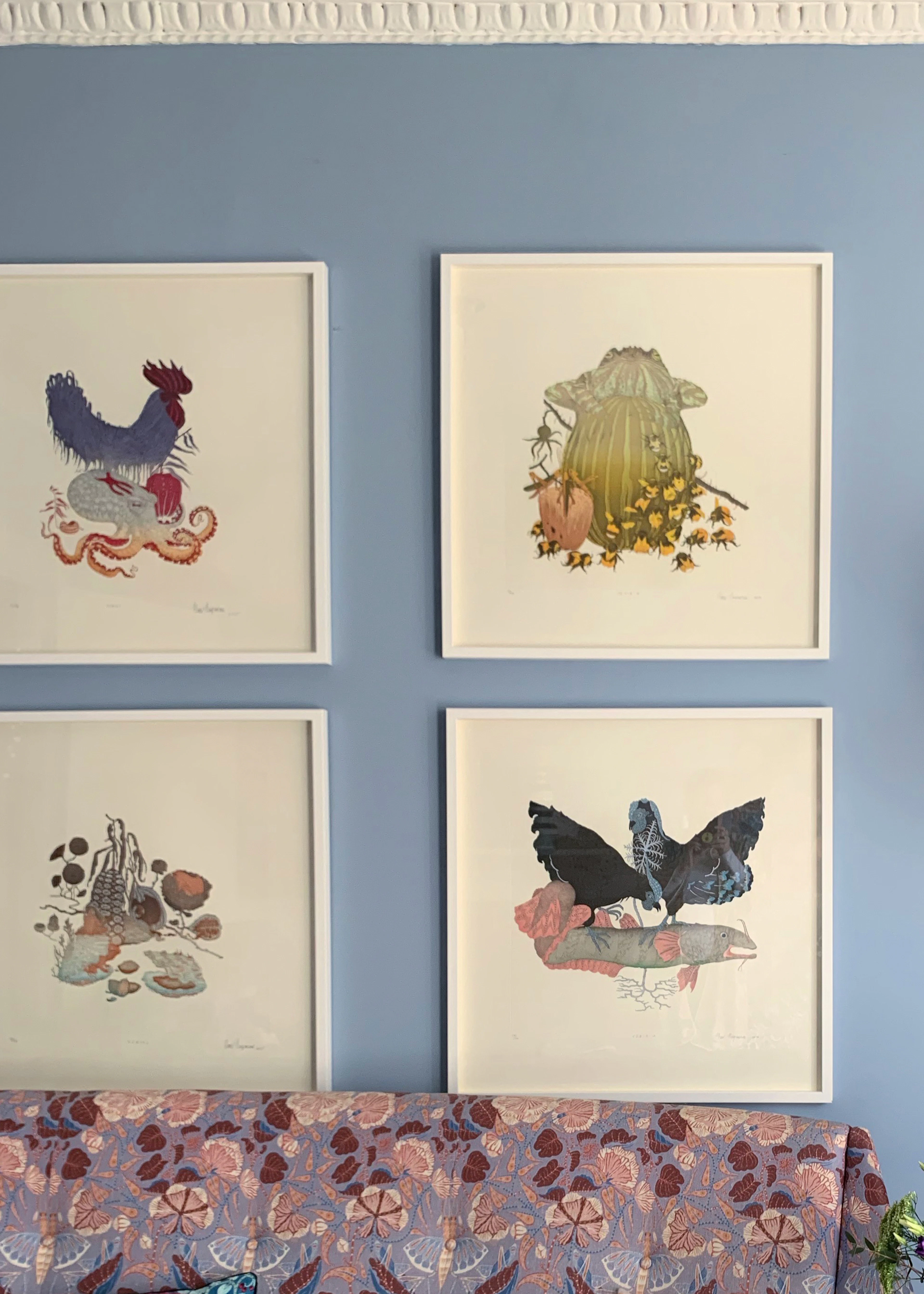 Gallery wall of four framed Klaus Haapaniemi art prints of birds and creatures on a blue wall above a patterned sofa