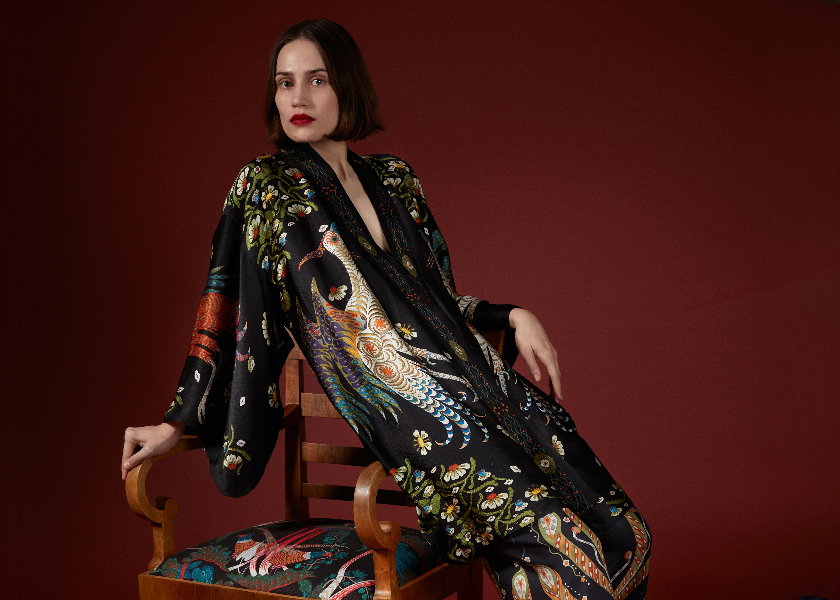 Woman in the black Firebird kimono seated on a wooden chair against a deep red wall