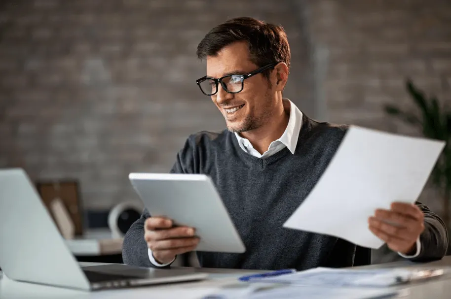 Homem sorrindo, usando óculos, trabalhando em escritório com notebook e tablet, enquanto analisa e segura folhas impressas com anotações, em ambiente corporativo.