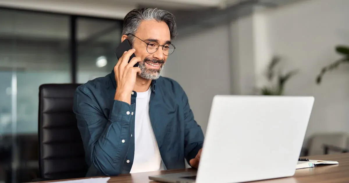 Homem com cabelo grisalho e barba fala ao telefone no escritório, enquanto utiliza um notebook aberto em sua mesa, em ambiente corporativo