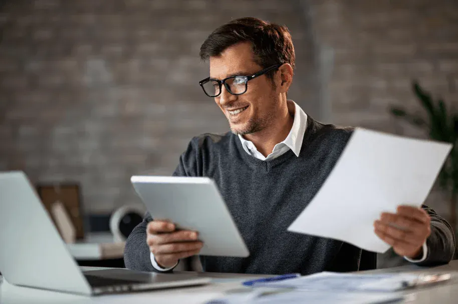 Homem usando óculos em home office sorri enquanto lê documentos e consulta um tablet e um laptop em uma mesa, com folhas e papel para trabalho